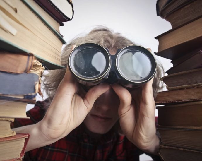 Young unrecognizable person sitting at table with stacked old books and looking through binoculars