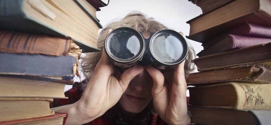 Young unrecognizable person sitting at table with stacked old books and looking through binoculars